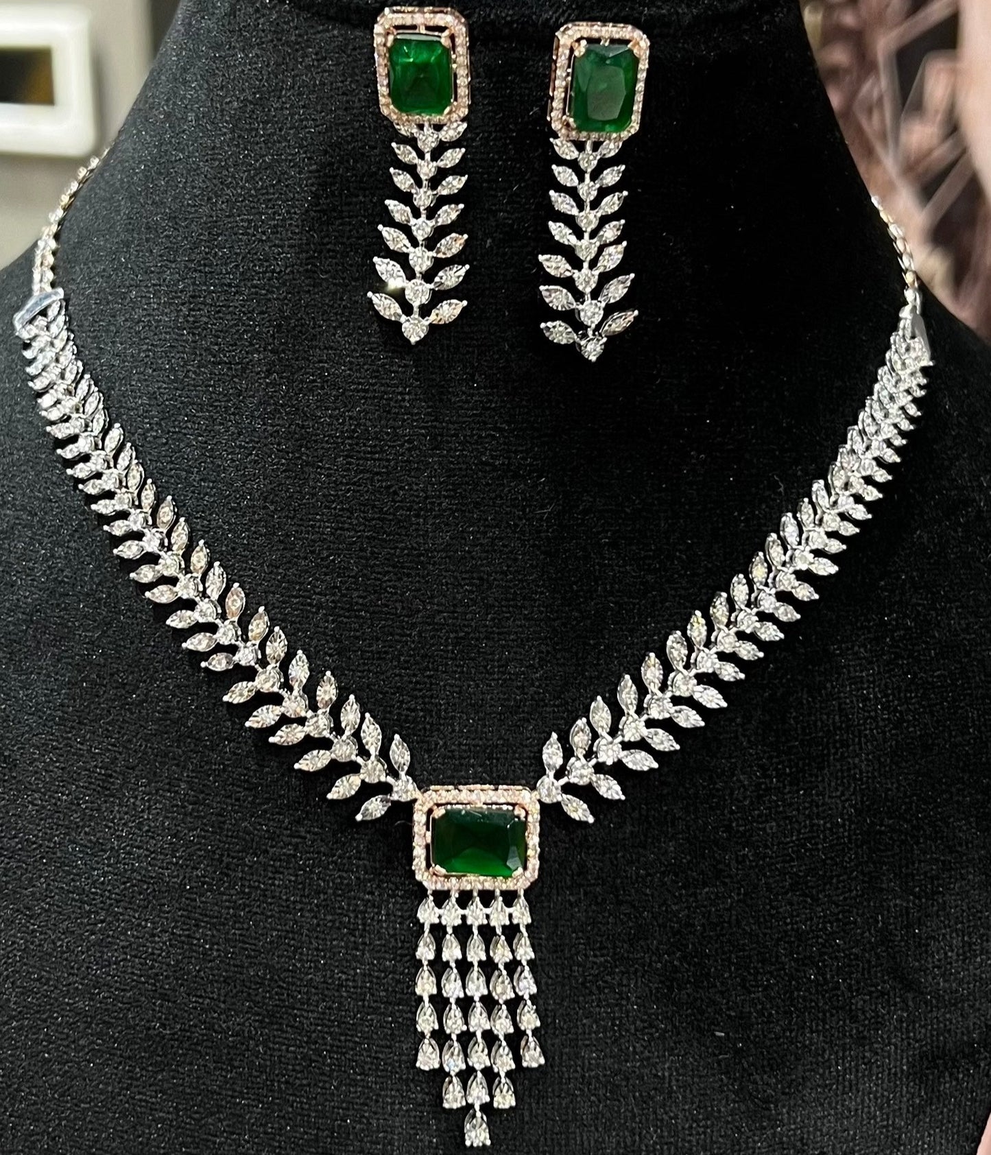 Diamond necklace set featuring a radiant green color stone centerpiece framed by diamond leaves and cascading diamond drops, paired with matching earrings on a mannequin bust.
