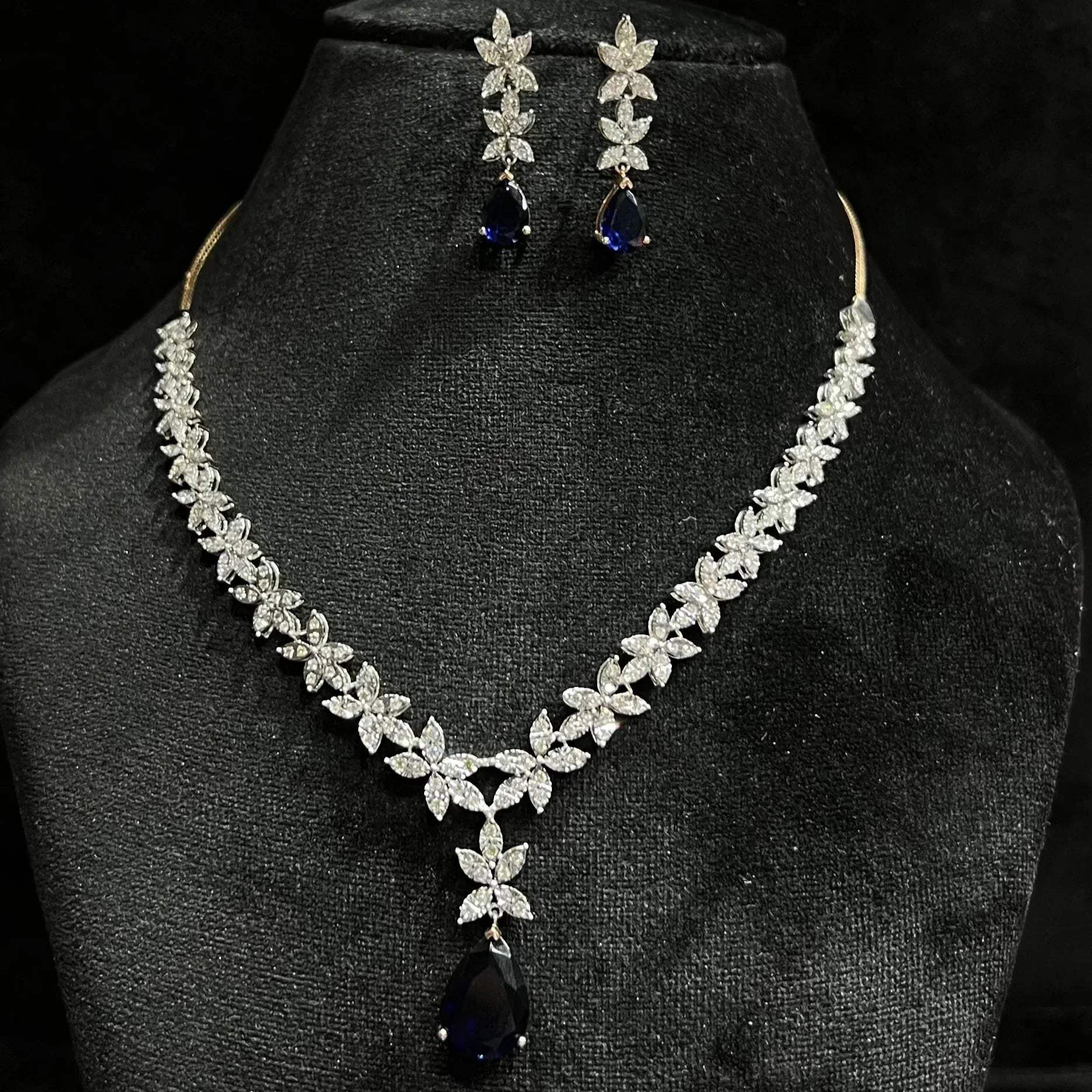 A gold diamond necklace set with matching earrings displayed on a mannequin bust, featuring a chain of gold and a series of graduated square and round cut diamonds & blue stones.