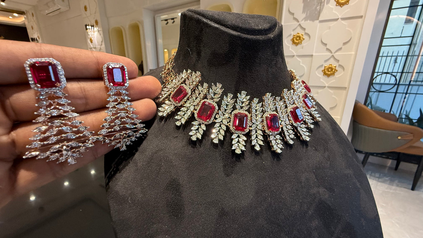 Diamond necklace set with emerald-cut color stones and leaf-shaped diamond clusters is displayed on a mannequin and matching color stone and diamond earrings are displayed on a hand.