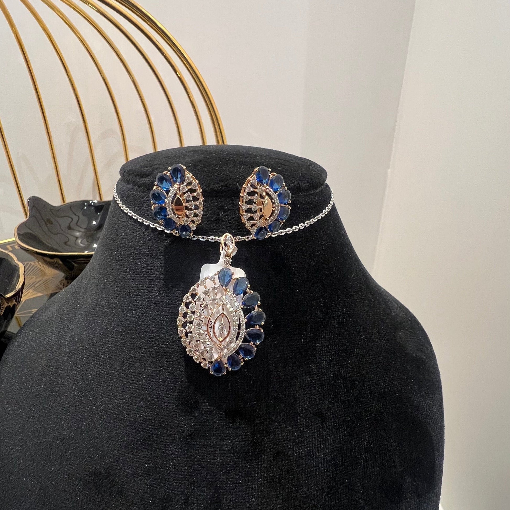 A gold diamond pendant with blue stone accompanied by matching earrings, displayed on a black mannequin bust.