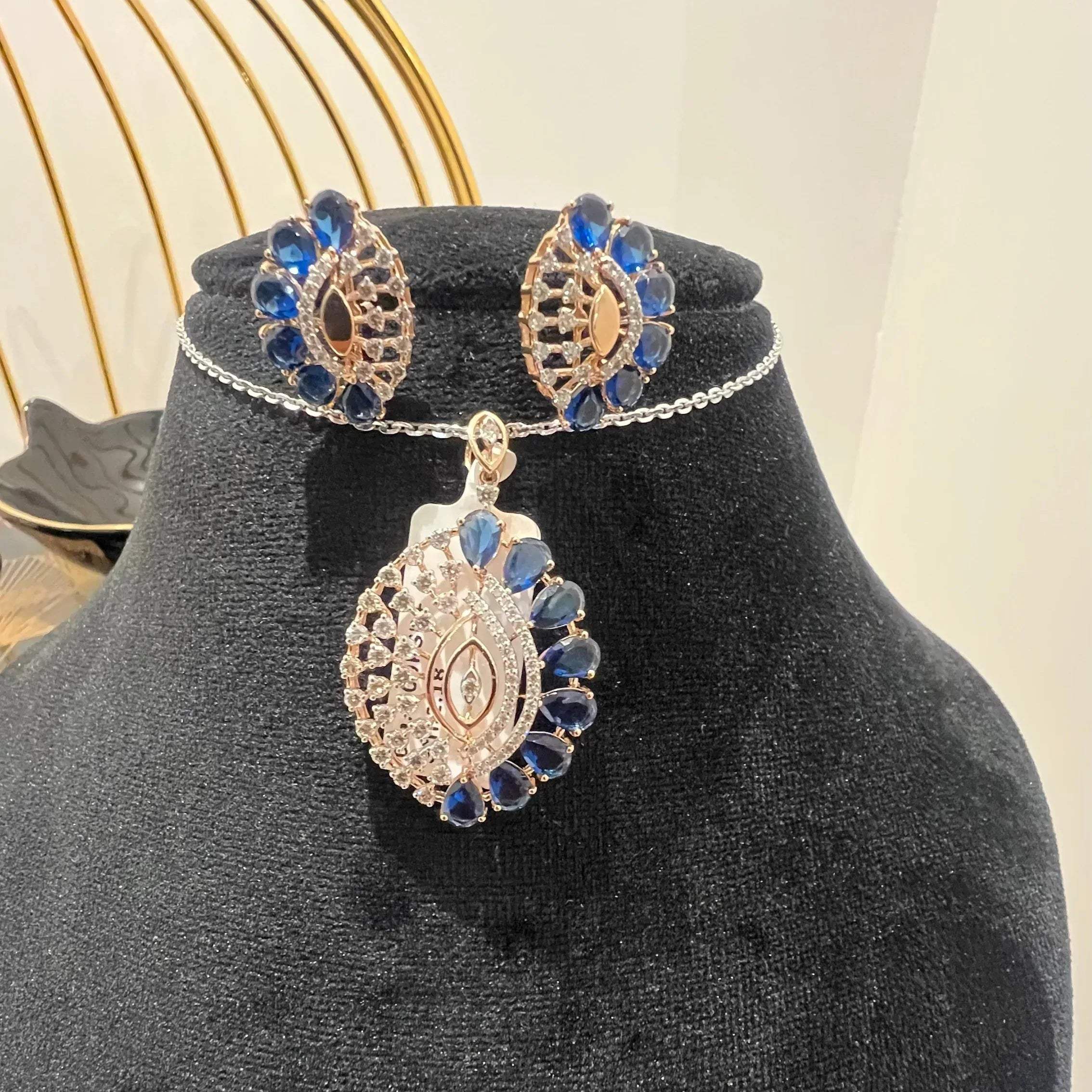 A gold diamond pendant with blue stone accompanied by matching earrings, displayed on a black mannequin bust.