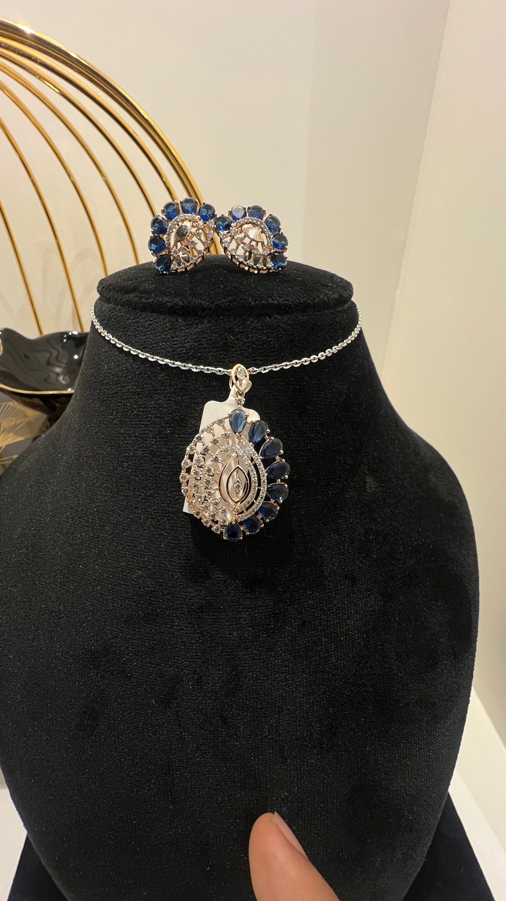A gold diamond pendant with blue stone accompanied by matching earrings, displayed on a black mannequin bust.