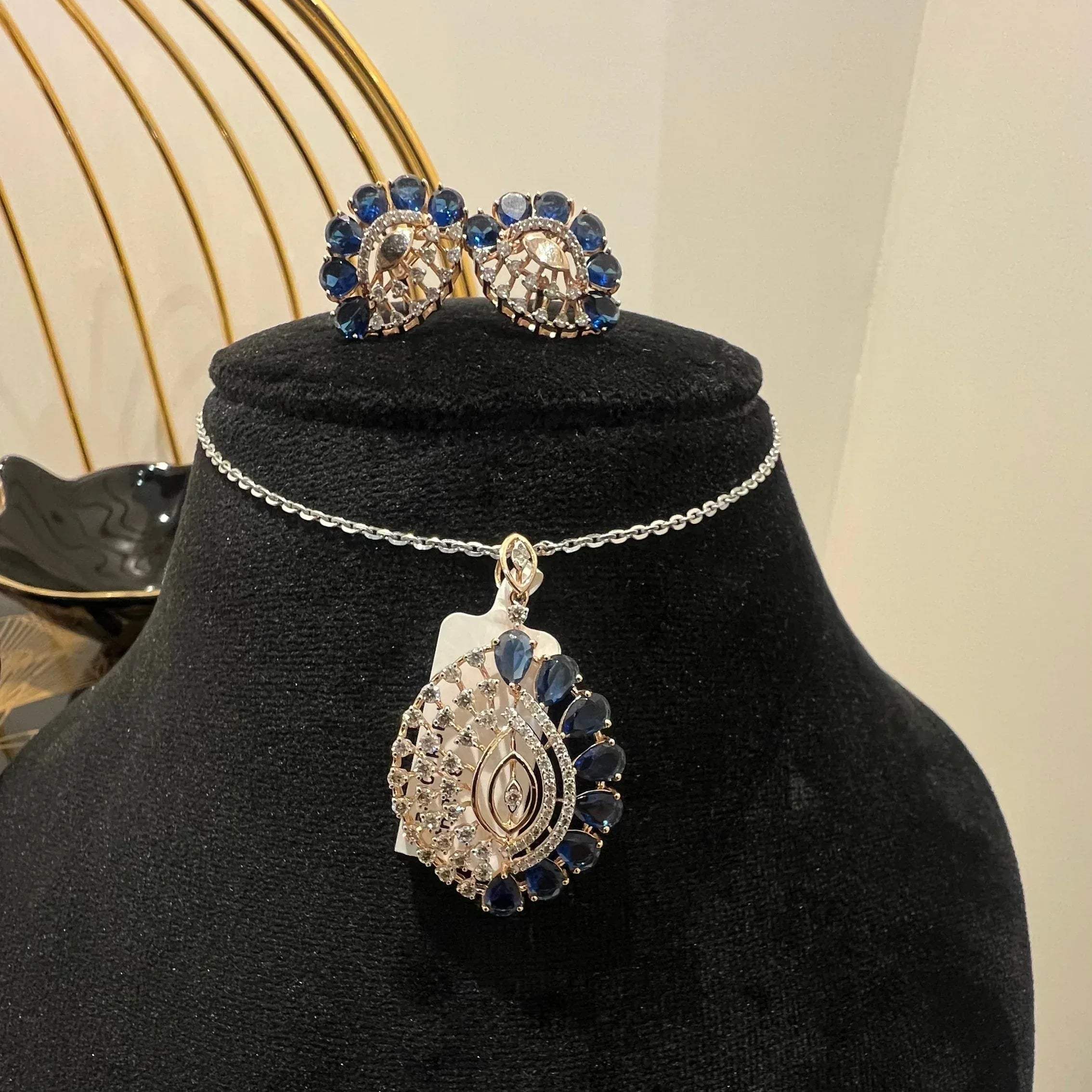 A gold diamond pendant with blue stone accompanied by matching earrings, displayed on a black mannequin bust.