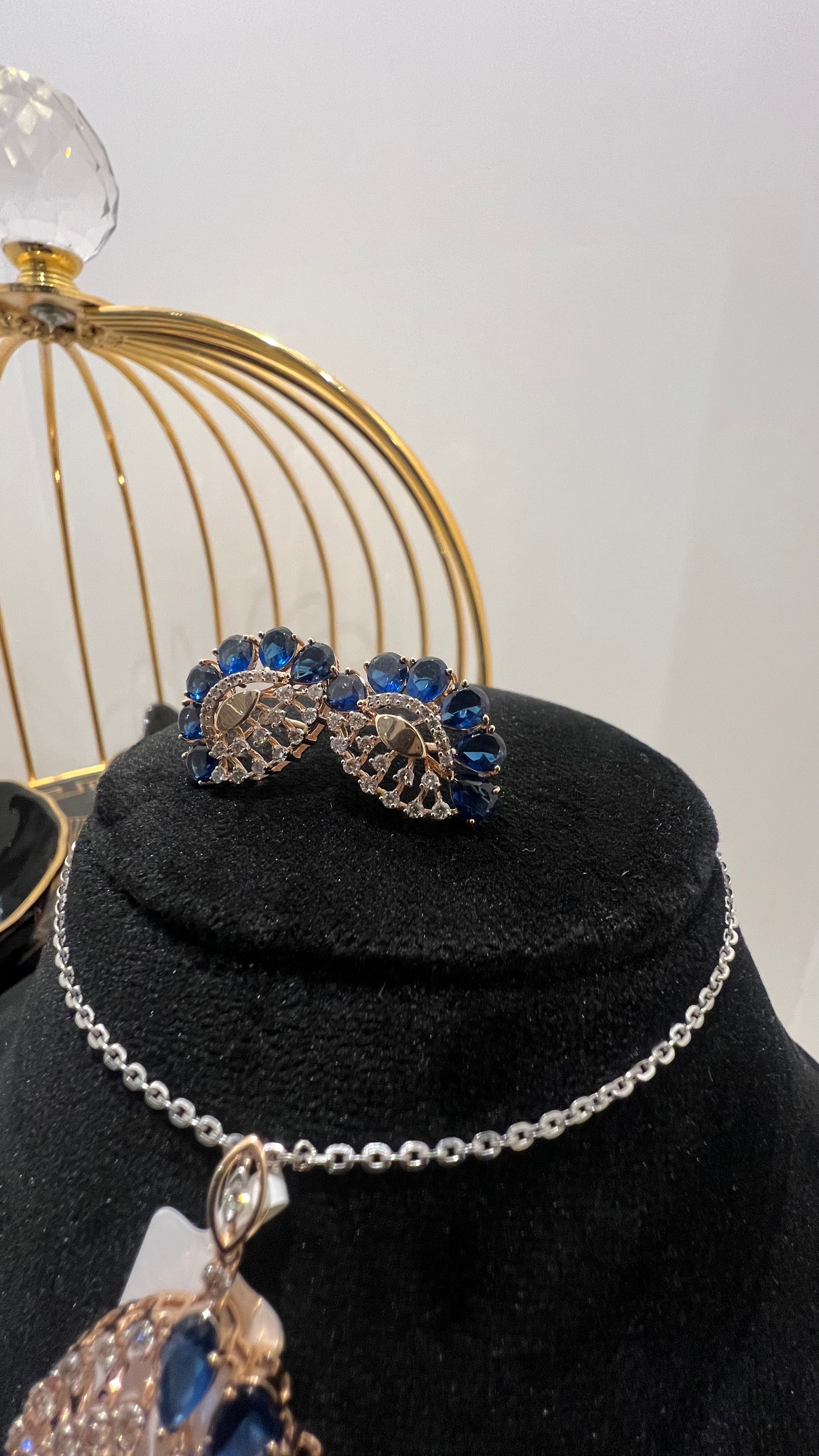 A gold diamond pendant with blue stone accompanied by matching earrings, displayed on a black mannequin bust.