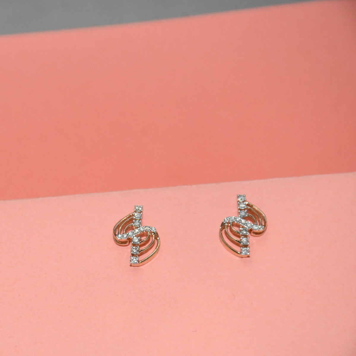 A pair of gold-toned spiral stud earrings adorned with small clear diamonds, displayed on a coral pink background.