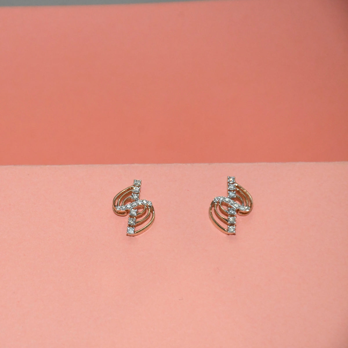 A pair of gold-toned stud earrings with small clear diamonds, displayed on a split pink and peach background.