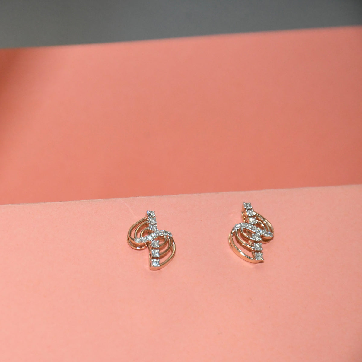 A pair of small gold-toned stud earrings with clear diamond accents, displayed on a peach-colored surface against a gray background.