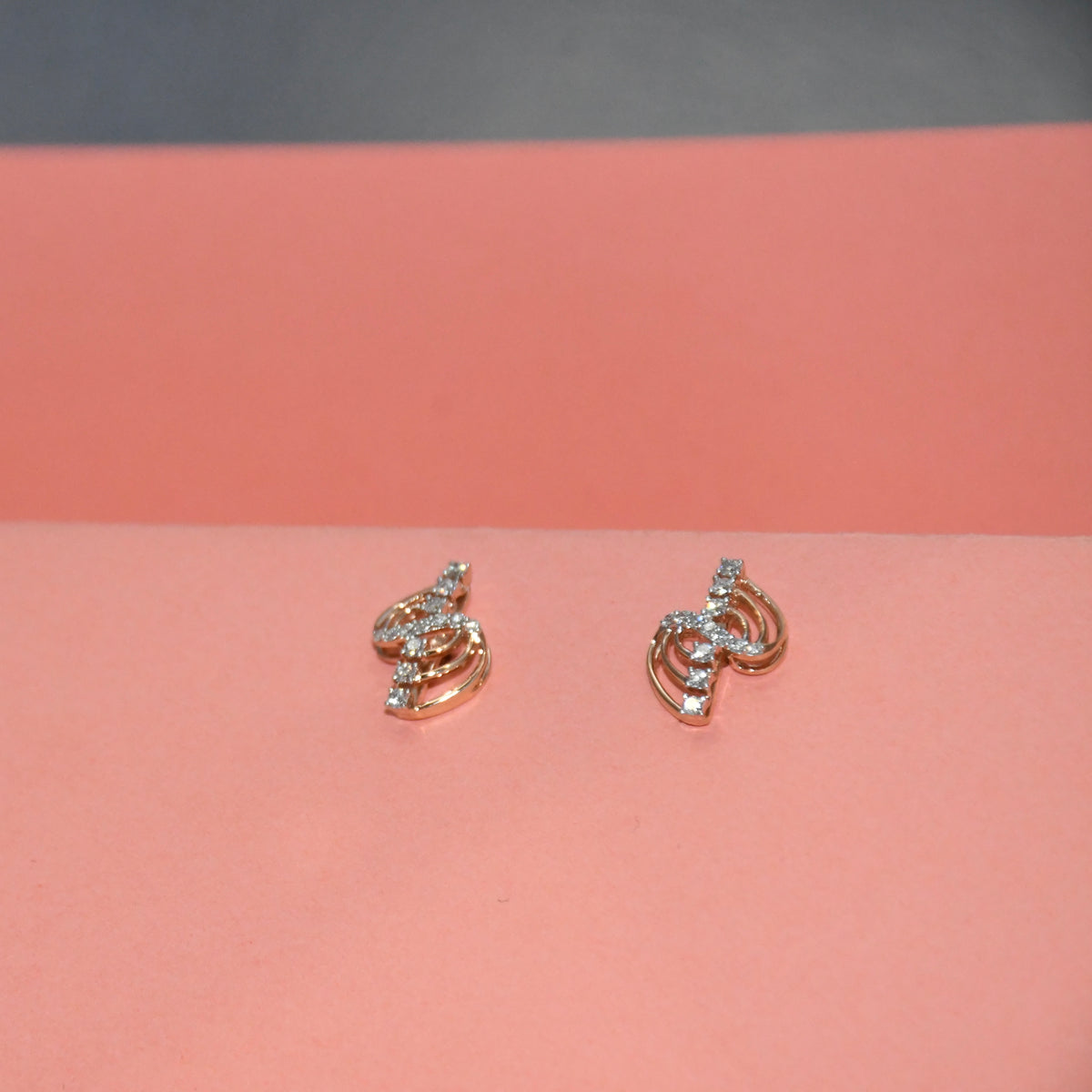 A pair of small gold earrings with clear diamonds, displayed on a pink surface against a dark gray background.
