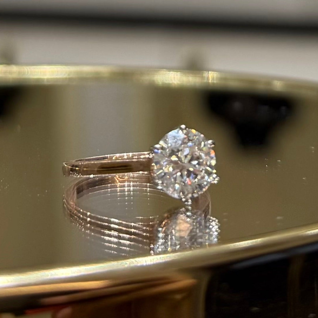A close-up of classic solitaire lab grown diamond ring with a round-cut diamond set in a four-prong gold band, displayed on a reflective surface.