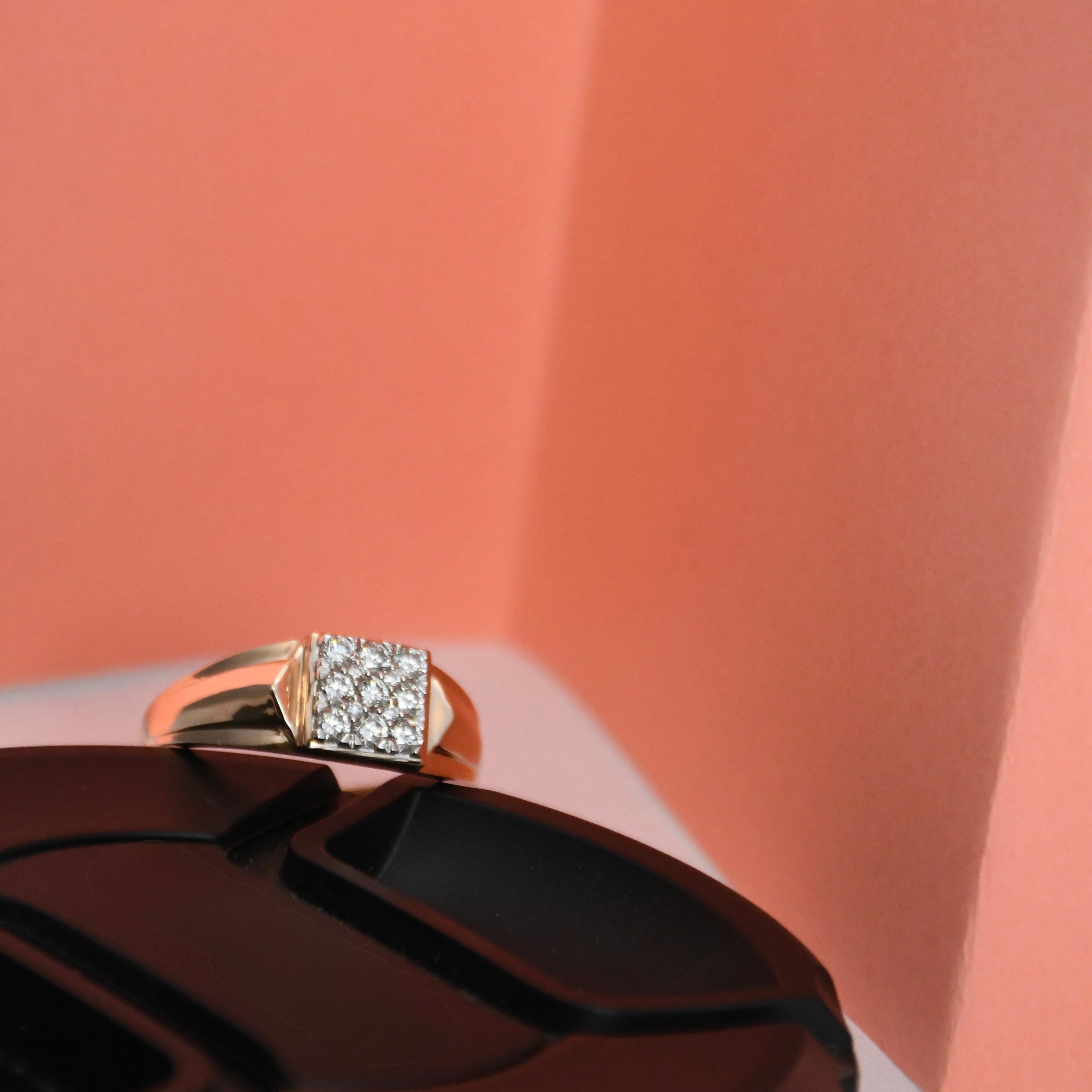 A gold ring with a square pavé diamond setting, displayed on a black surface against a peach-colored background.