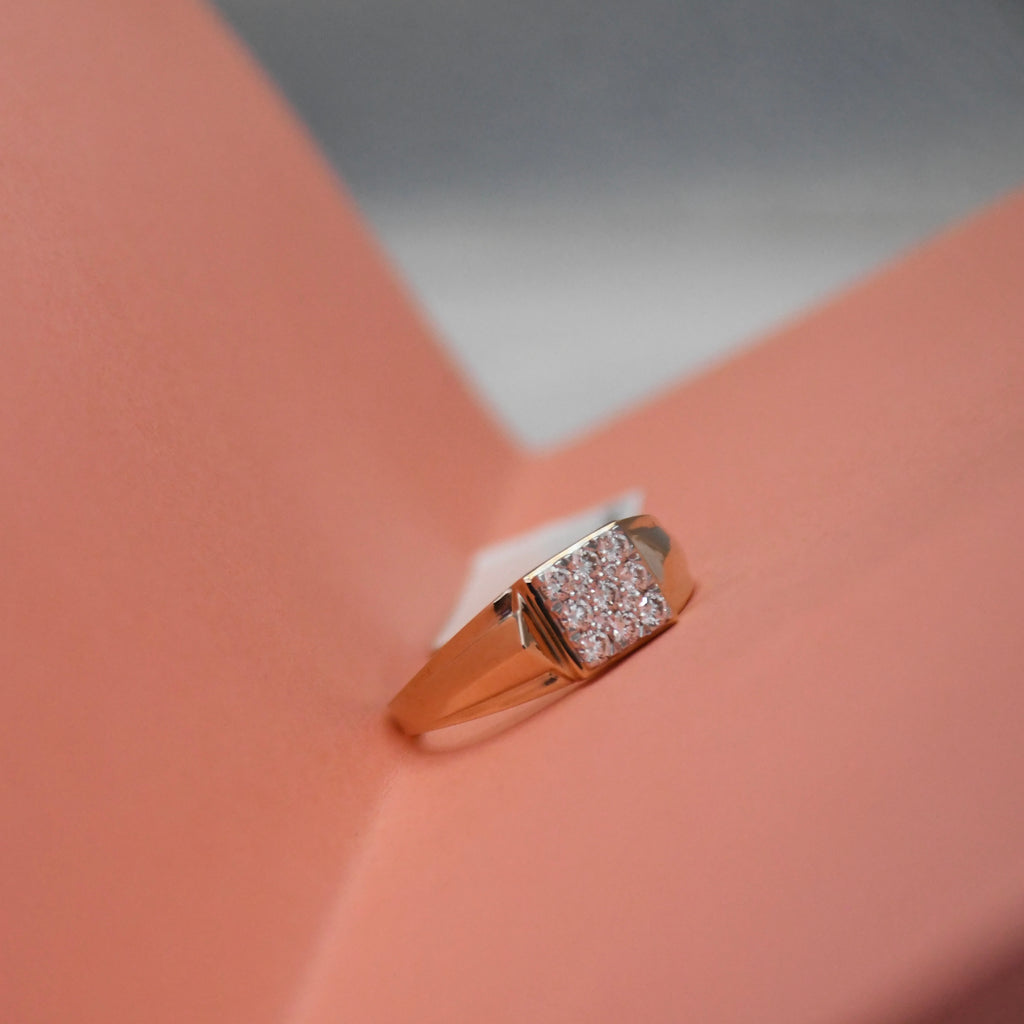A rose gold signet ring with a square pavé diamond setting, displayed on a peach-colored geometric surface.
