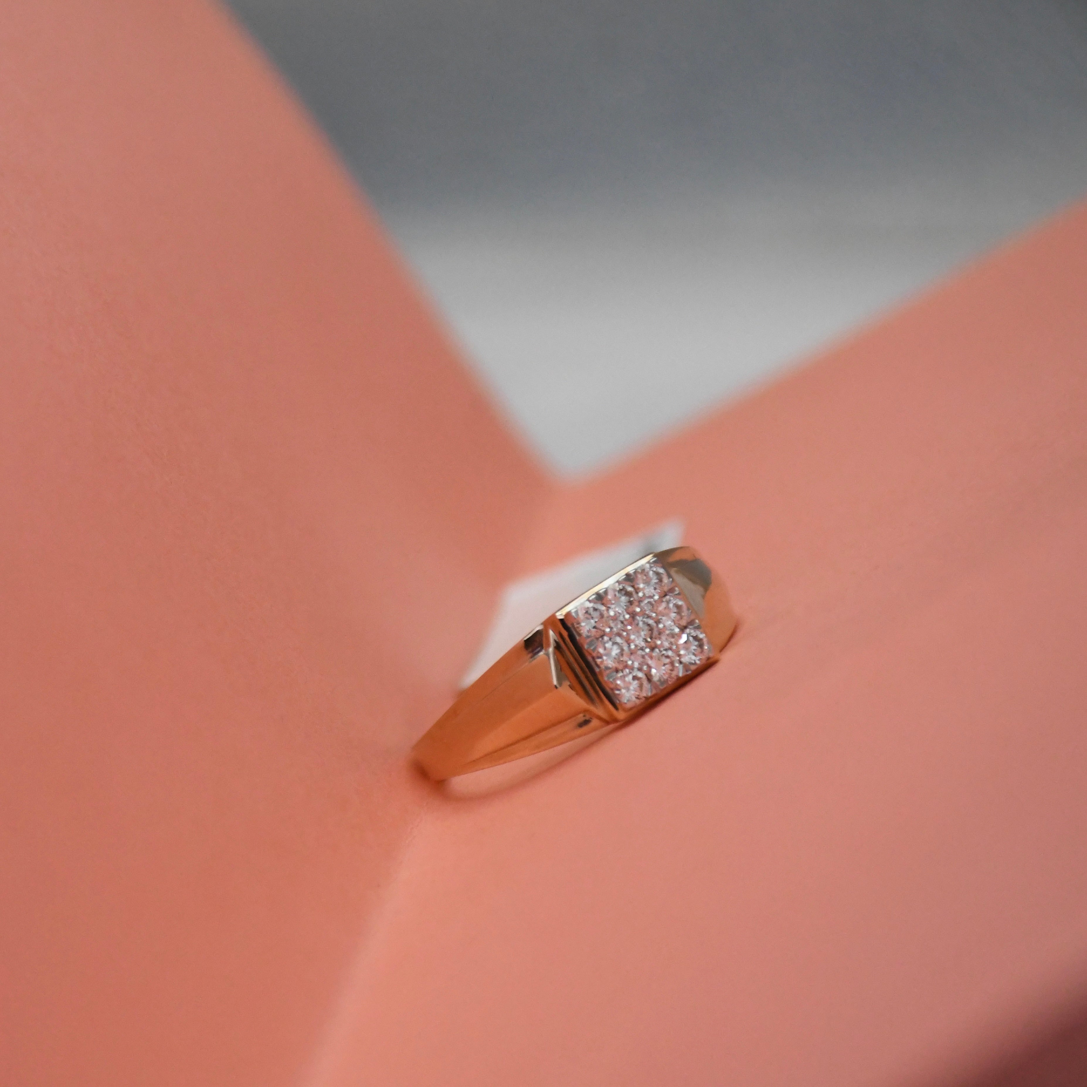 A rose gold signet ring with a square pavé diamond setting, displayed on a peach-colored geometric surface.