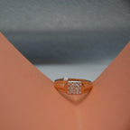 A rose gold ring with a square pavé diamond setting, displayed on a peach-colored ring stand against a gray textured background.