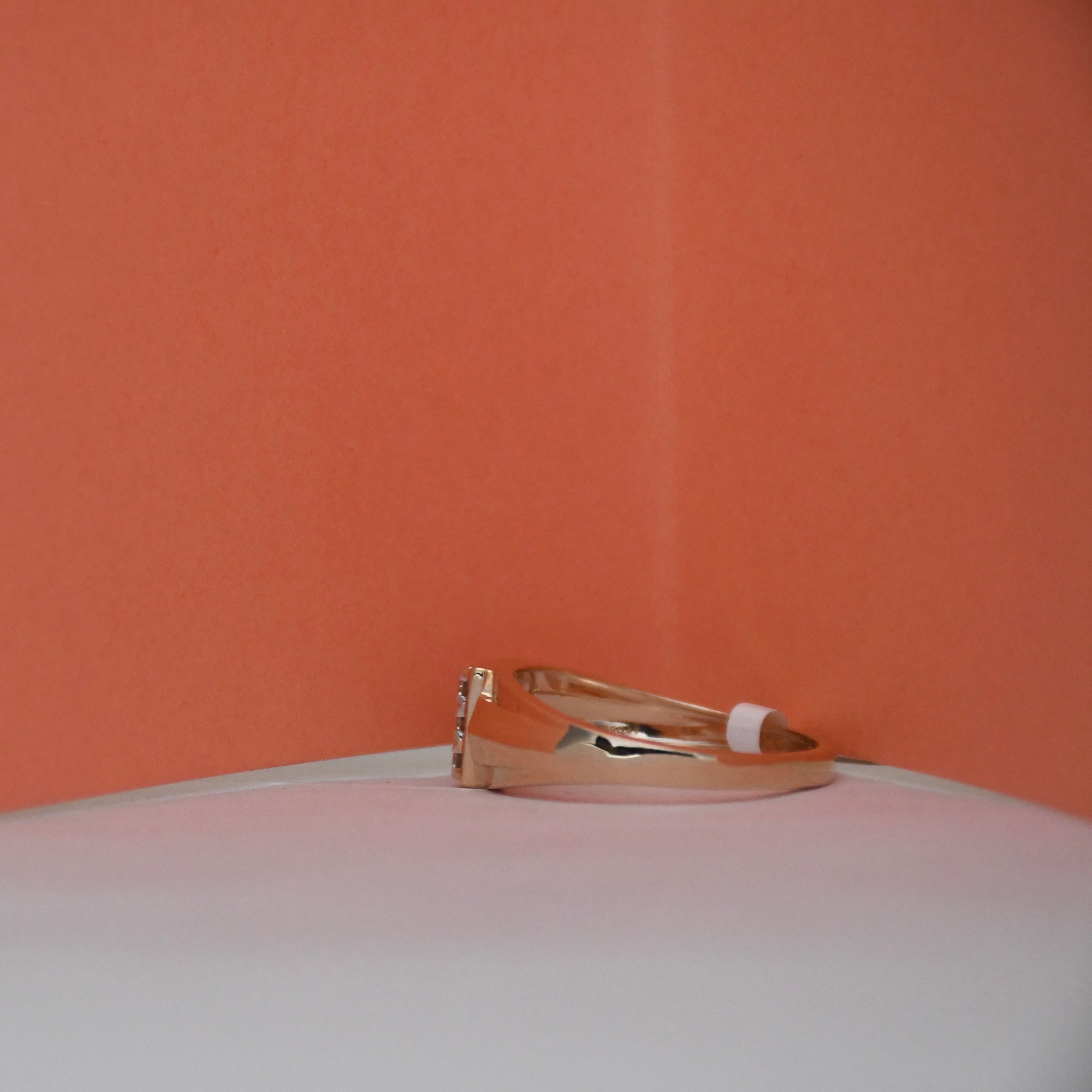 A rose gold ring with a small clear gemstone on the band, resting on a white surface against a textured orange background.