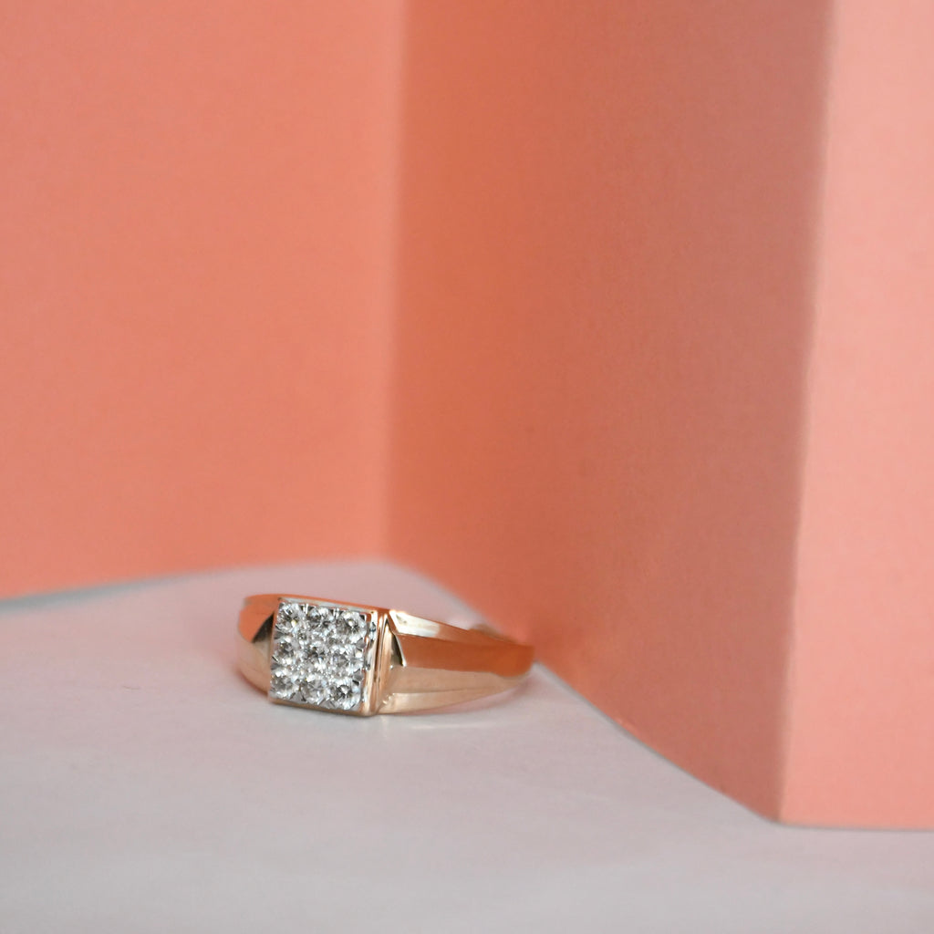 A rose gold ring with a square cluster of clear diamonds, resting on a white surface against a peach-colored background.