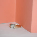 A rose gold ring with a square cluster of clear diamonds, resting on a white surface against a peach-colored background.