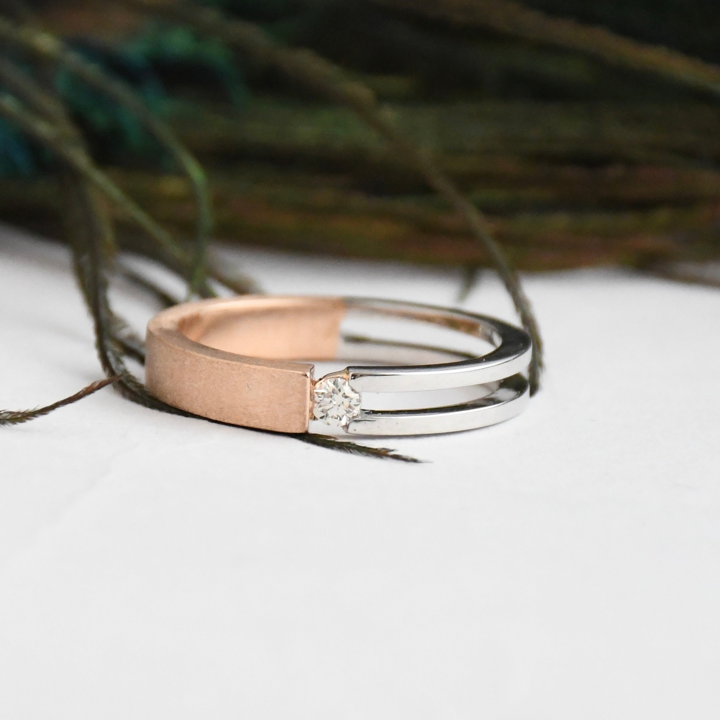 Two tone gold solitaire diamond ring, one rose gold and one white gold, on a white surface with a blurred natural background.