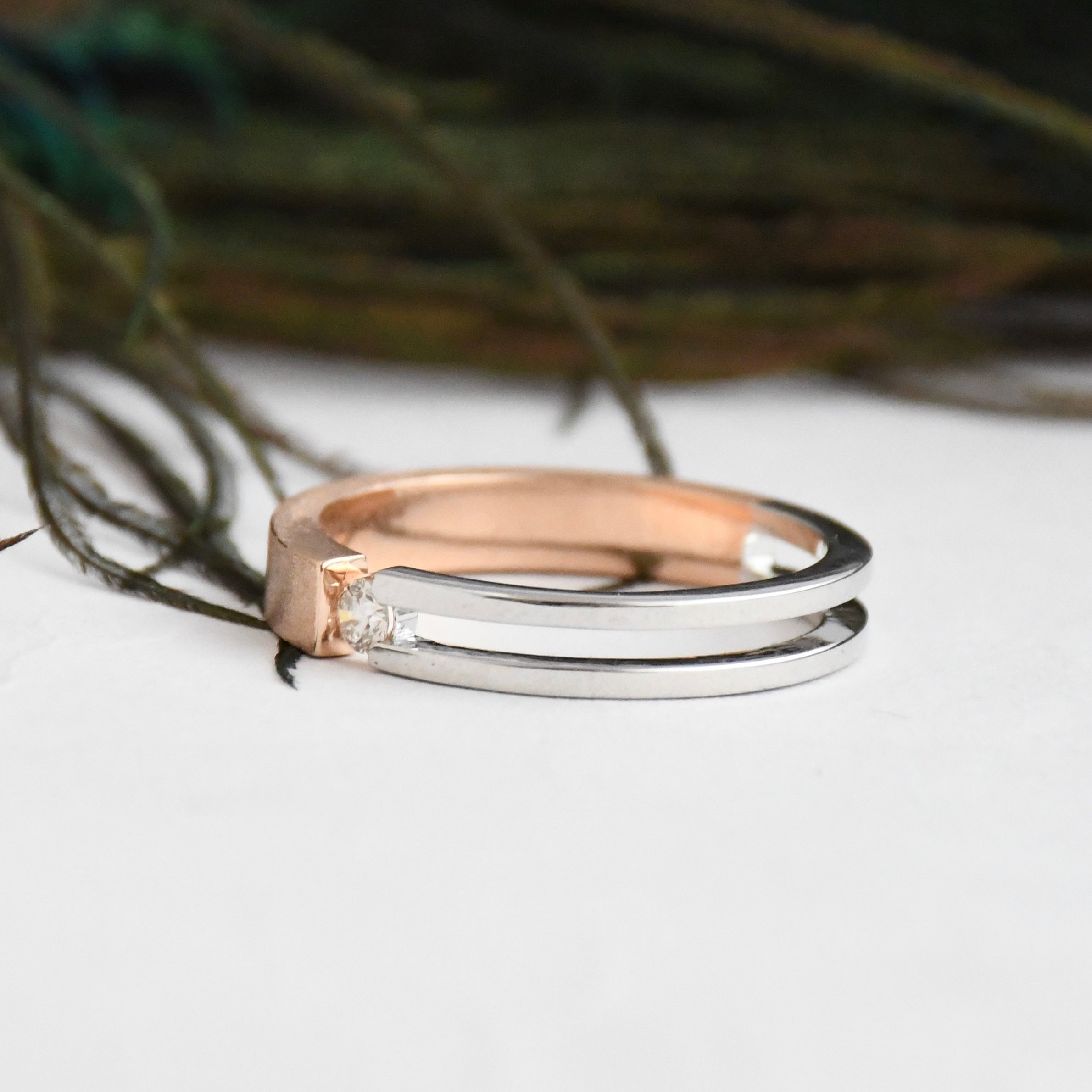 Two tone gold solitaire diamond ring, one rose gold and one white gold, on a white surface with a blurred natural background.