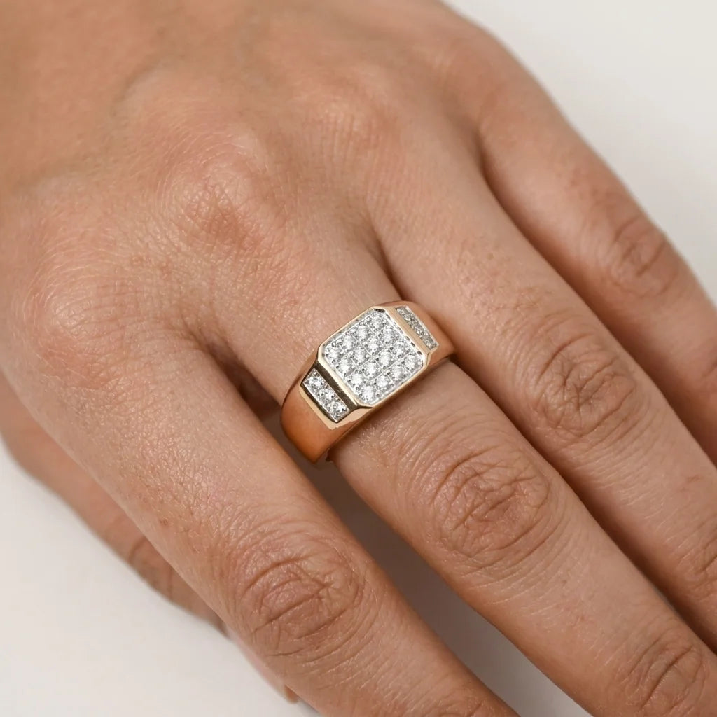 A gold ring with a square pavé diamond center and a small side channel of diamonds, displayed on a person's hand against a white surface.