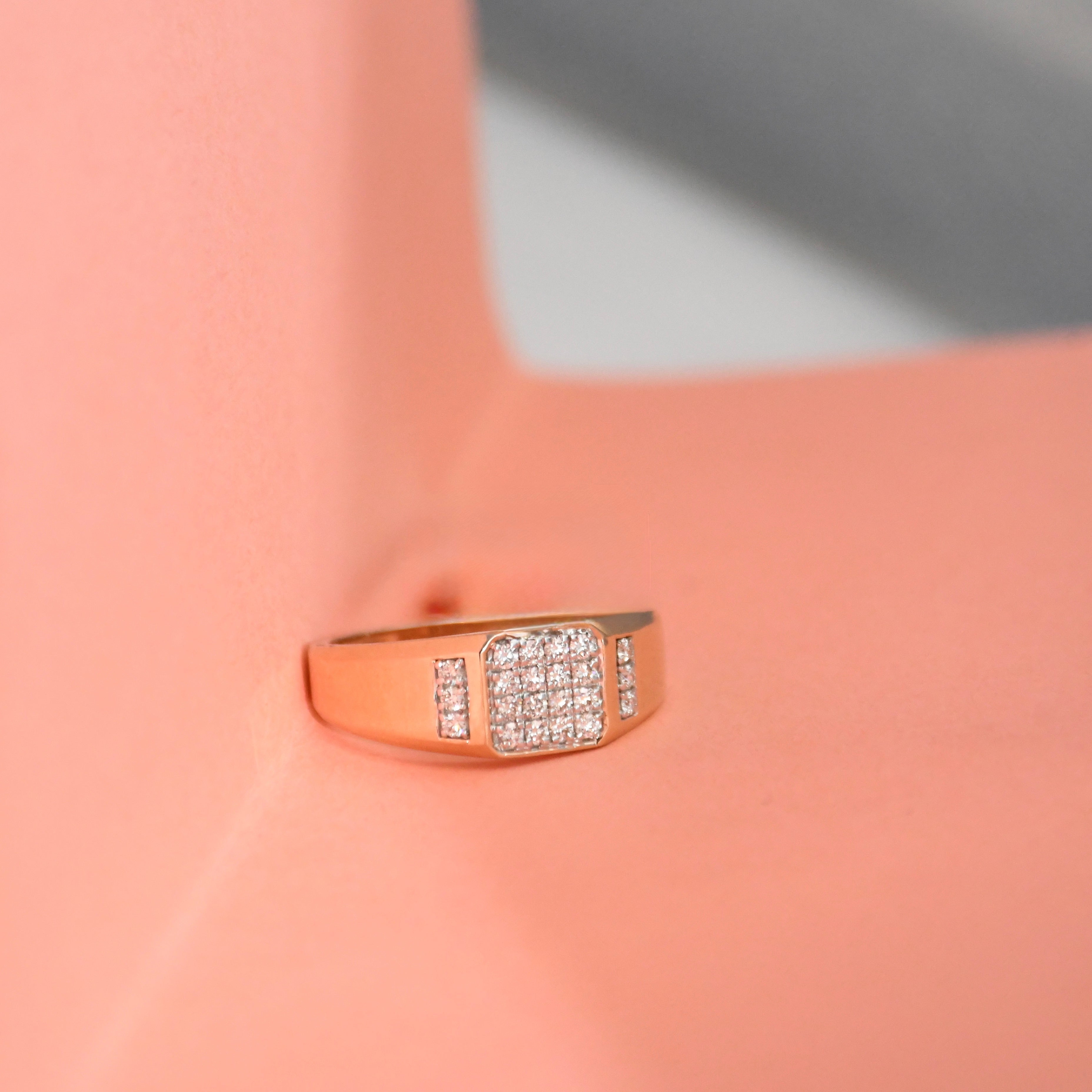 A rose gold signet-style ring with a square pavé diamond center and smaller diamond accents on the shoulders, displayed on a peach-colored jewelry stand.