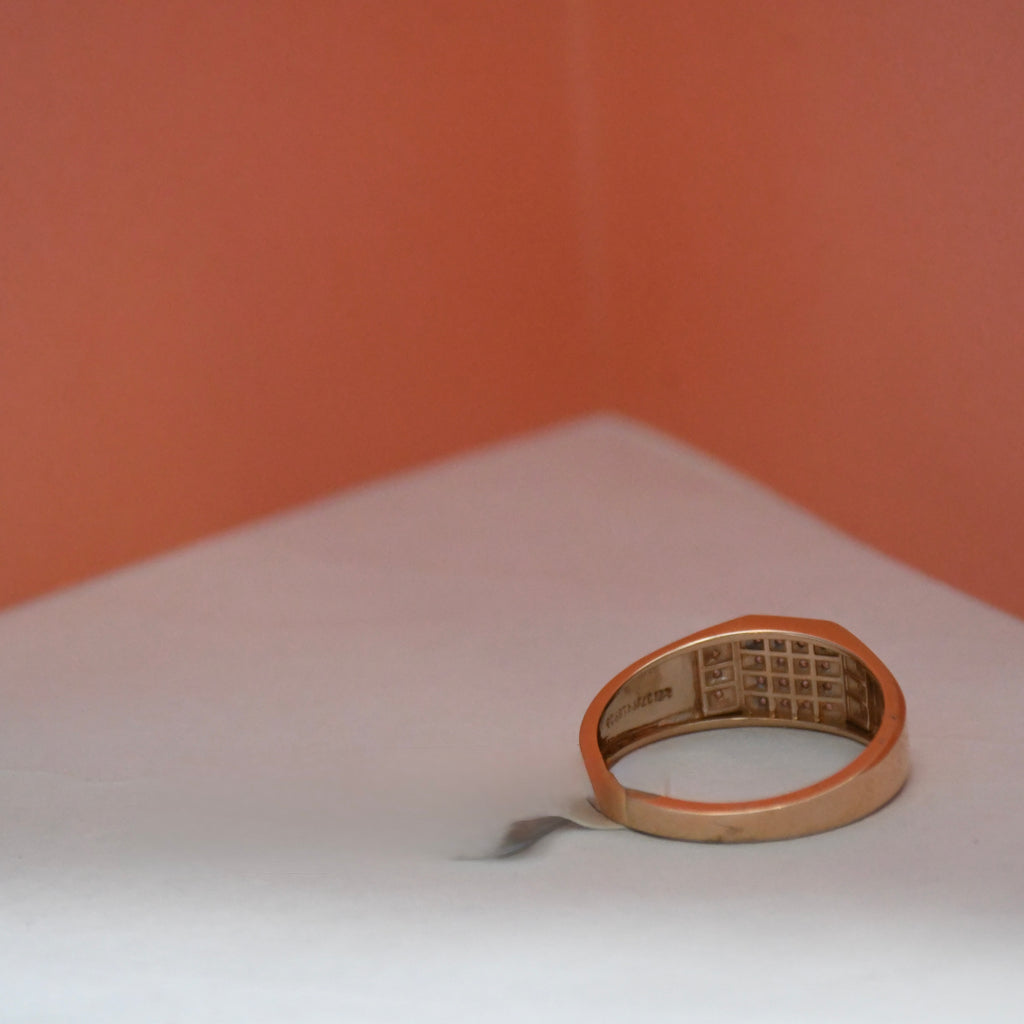 A gold signet ring with a grid-patterned center, resting on a white surface against a warm peach-colored background.