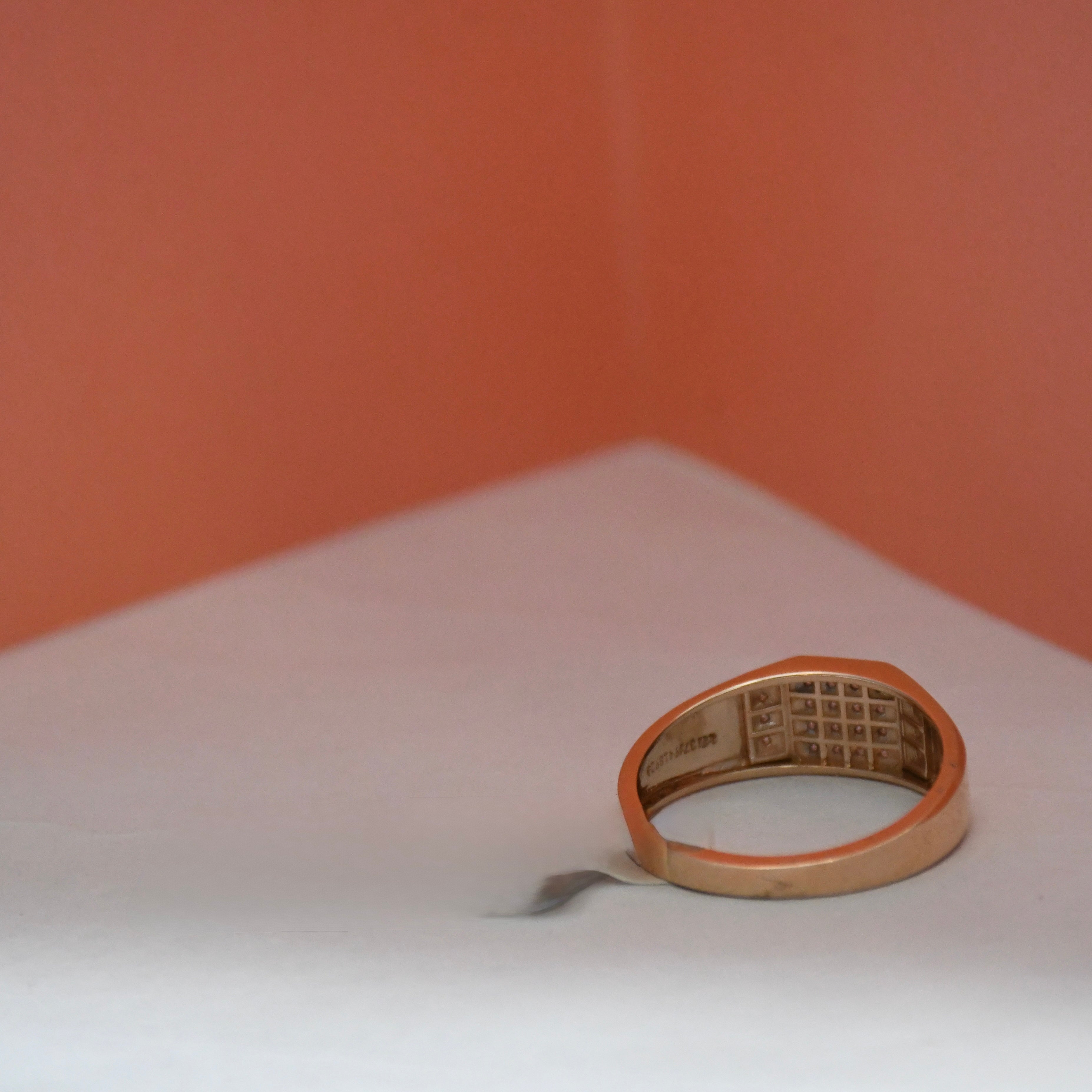 A gold signet ring with a grid-patterned center, resting on a white surface against a warm peach-colored background.