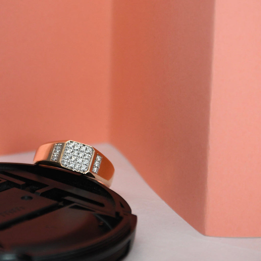 A rose gold ring with a square pavé diamond center and side accents, resting on a black circular display stand against a peach and white background.