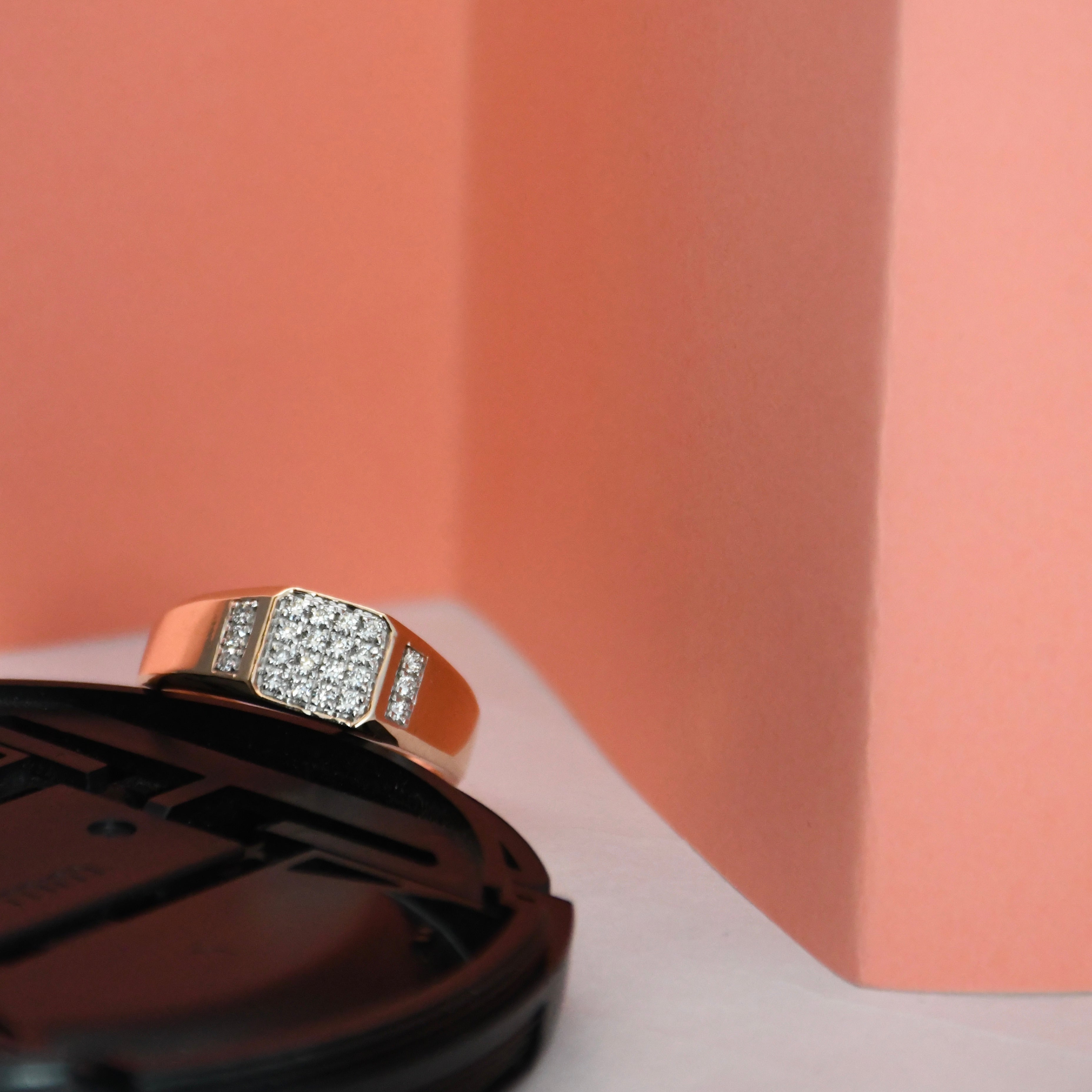 A rose gold ring with a square pavé diamond center and side accents, resting on a black circular display stand against a peach and white background.