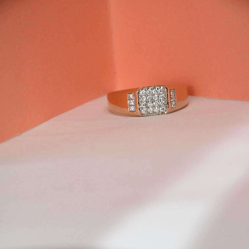 A rose gold ring with a square pavé diamond center and smaller diamond accents on the band, displayed on a white surface against a peach-colored background.