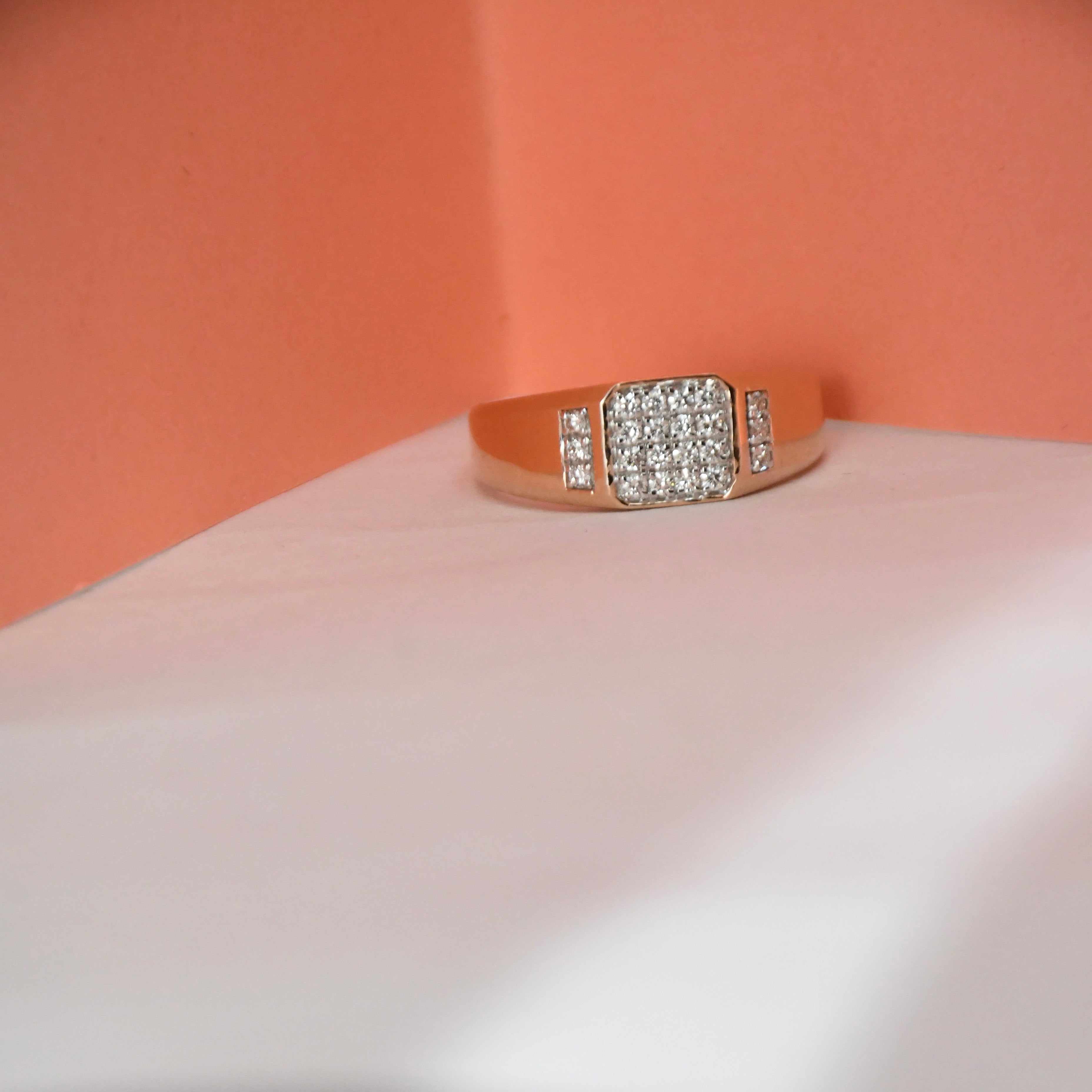 A rose gold ring with a square pavé diamond center and smaller diamond accents on the band, displayed on a white surface against a peach-colored background.