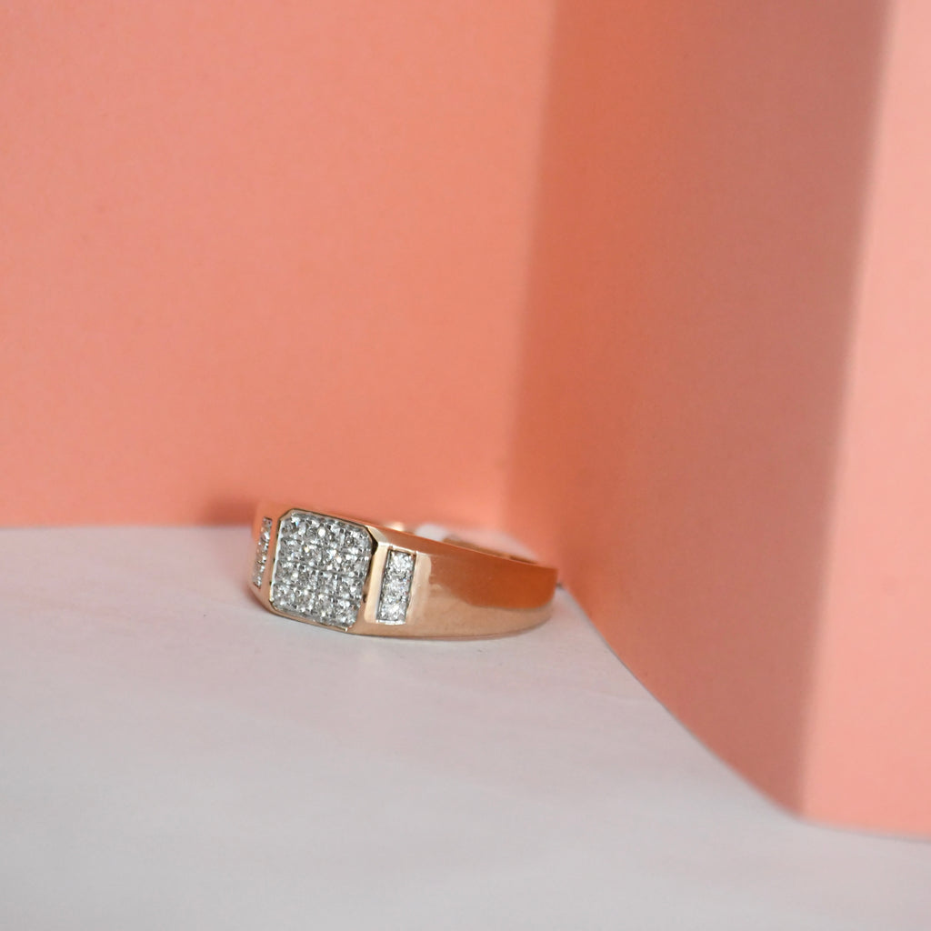 A gold ring with a square pavé diamond center and a small side channel of diamonds, displayed on a white surface against a peach-colored background.