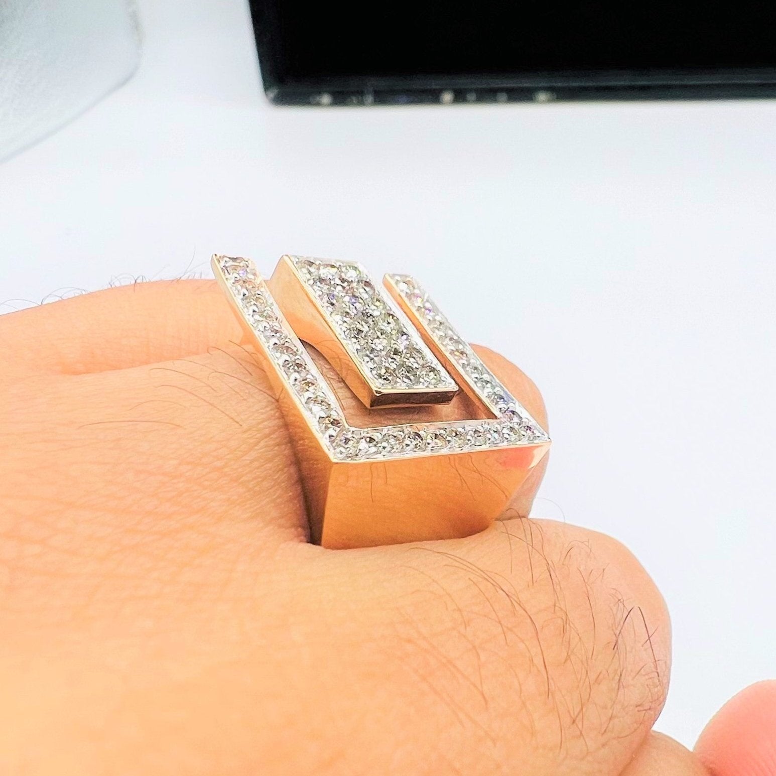 A man's hand displaying a gold diamond ring with a large square shape with round cut diamonds on top.