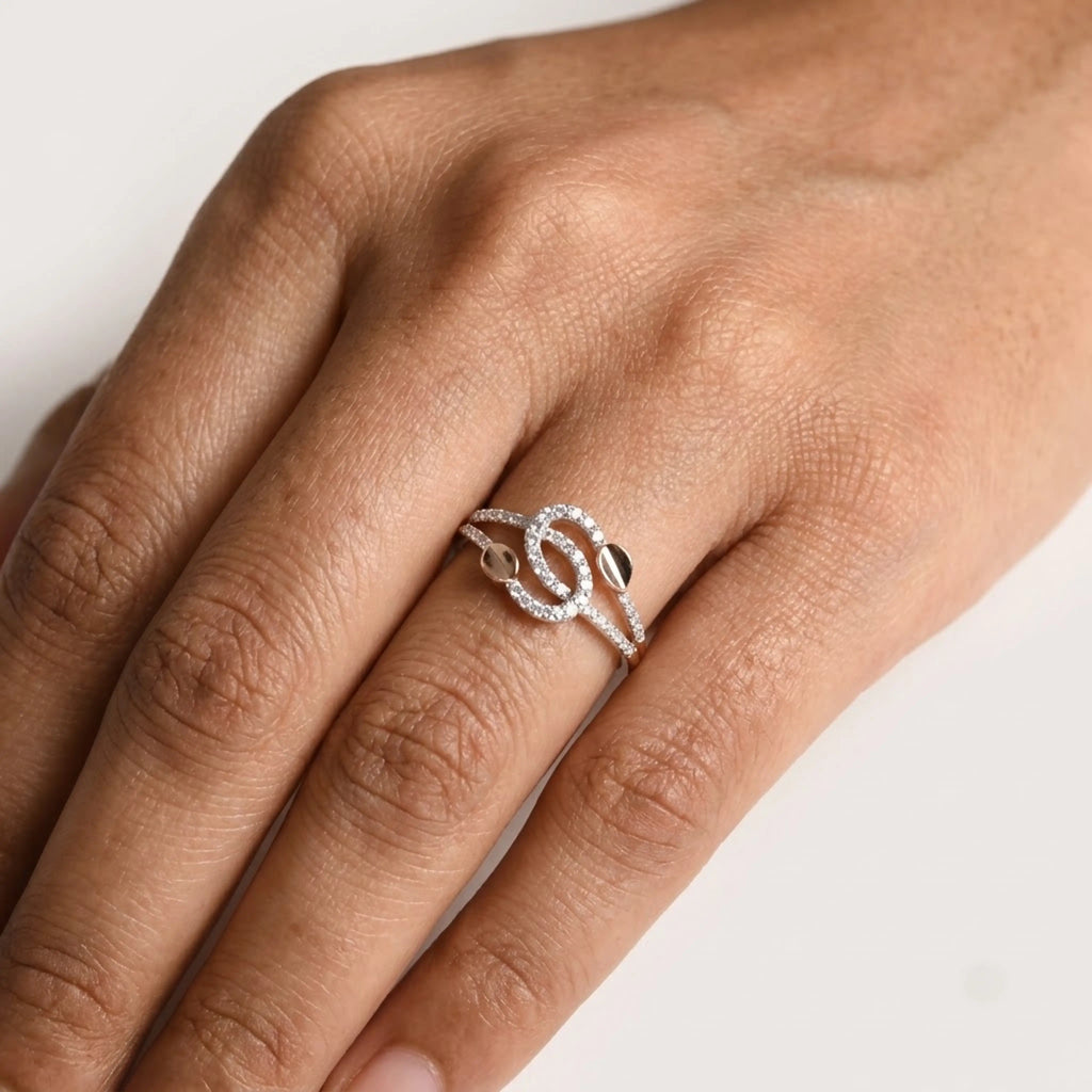 A gold toned ring with a double knot design and small clear stones, shown worn on a person's finger against a plain light background.