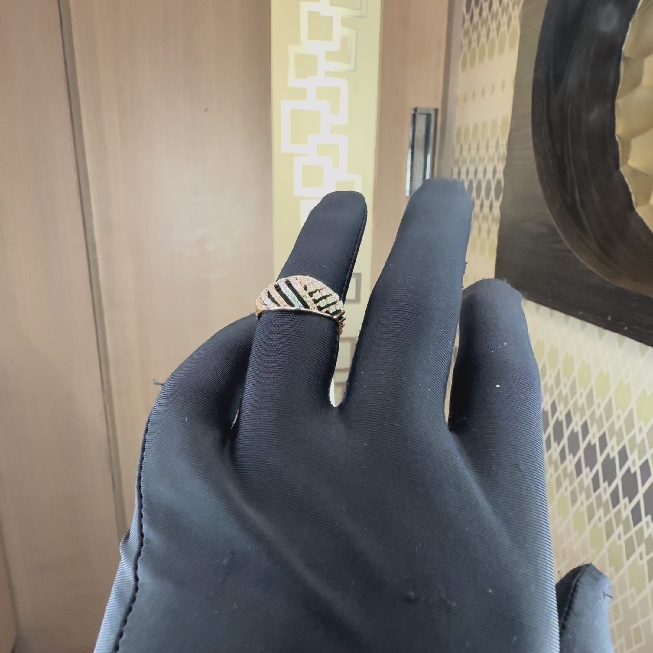 A rose gold ring featuring a curved, leaf-like design paved with small clear diamonds, worn by a hand which also worn black glove. The hand is tilting right and left to show the every angle of the ring.