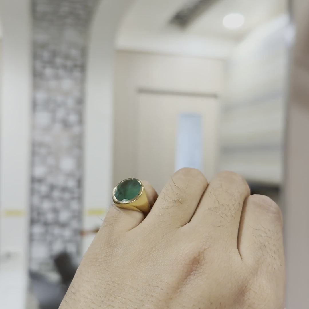 A person's hand is showing from the close which is worn a gold ring with a green emerald stone in its index finger and showing it from every angle by tilting the hand both sides against a neutral background.