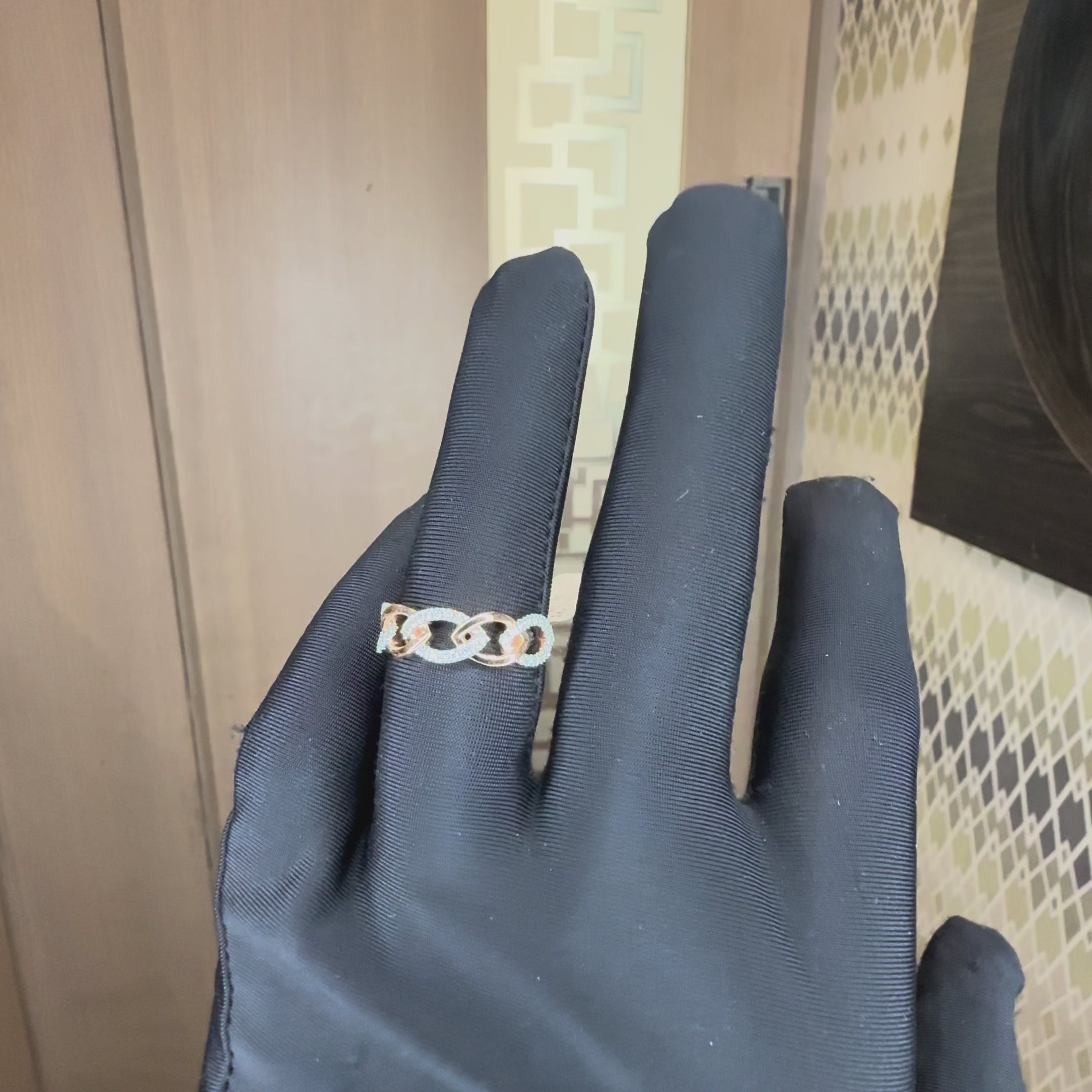 A close-up of a hand with black gloves wearing a delicate rose gold ring with small clear stones, set against a neutral background, showing the ring from every angle.