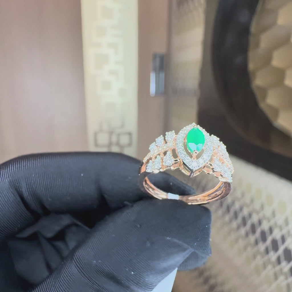 A rose gold ring featuring a central marquise-cut green emerald surrounded by a halo of small clear stones, with additional clear stones along the band, showing by a hand who is worn black gloves and tilting the ring both sides to show the every angle of the ring