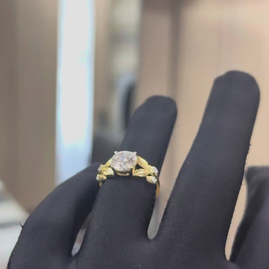 Gold ring with lab grown diamond and leaf designs against a natural background shown by a hand.