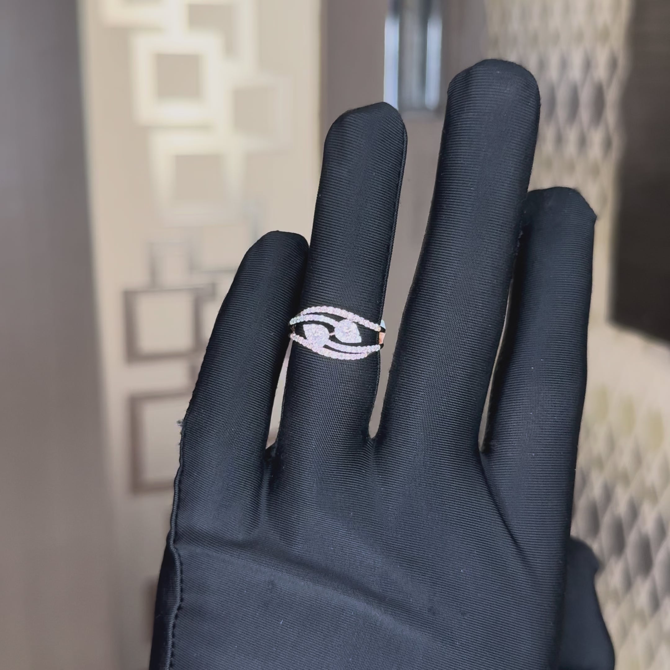 A rose gold ring with pavé-set clear stones, featuring a central pear shape cluster and four curved bands, worn in a hand's finger, which is also worn black gloves. The hand is tilting left and right to show the ring from every angle.