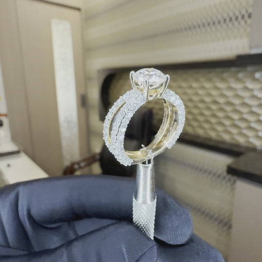 Gold ring with one solitaire diamond and small pave set diamonds is showing against a neutral background. Ring is holded by a ring holder by a person's hand.