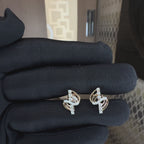 A pair of gold and diamond stud earrings with a curved, layered design, hold by a hand which is worn black glove and showing earrings from the close to show its every small detail against a neutral background.