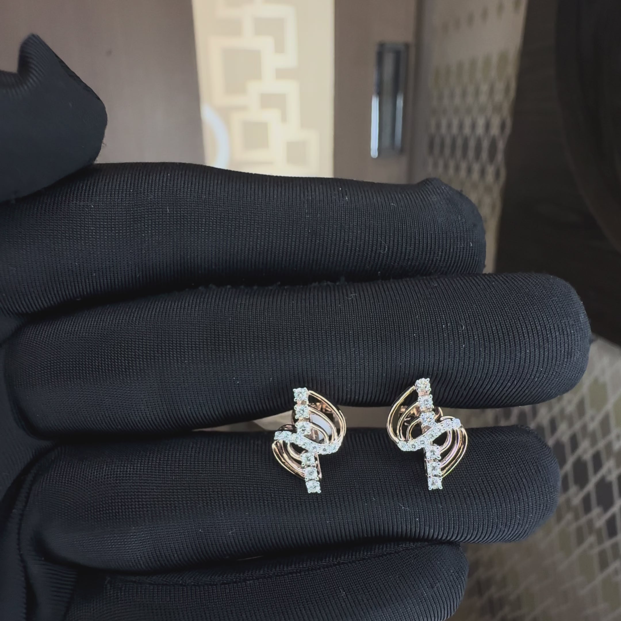 A pair of gold and diamond stud earrings with a curved, layered design, hold by a hand which is worn black glove and showing earrings from the close to show its every small detail against a neutral background.