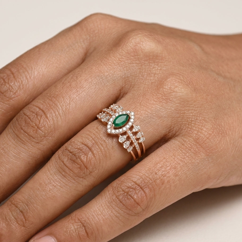A close-up of a hand wearing a rose gold ring with a marquise-cut green emerald center stone and surrounding pavé clear stones.
