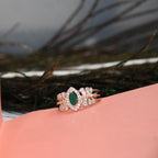 A rose gold ring with a central green marquise-cut gemstone and surrounding clear stones, displayed on a pink surface with a blurred dark background.