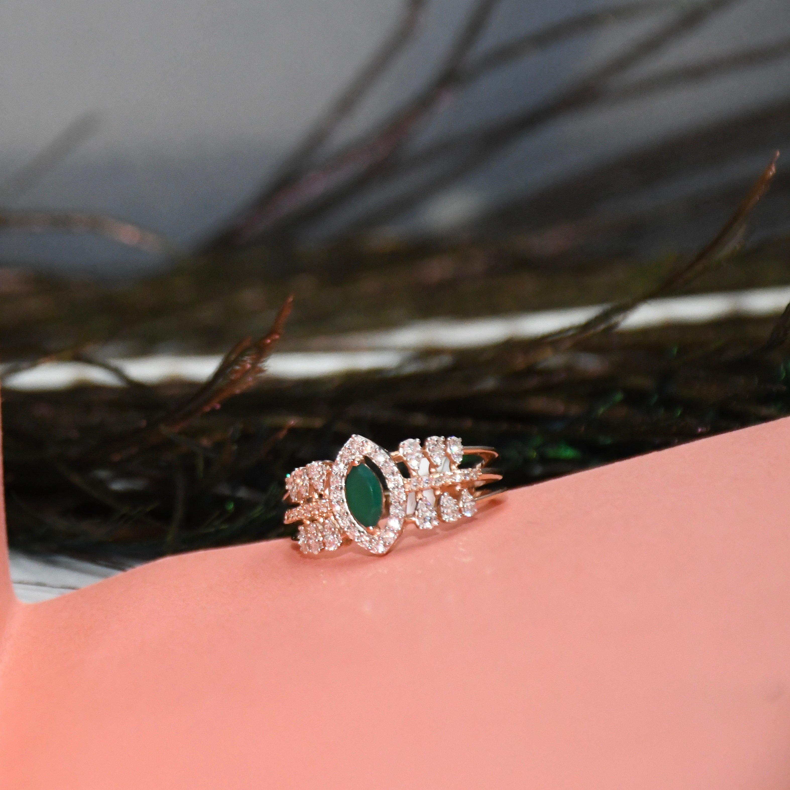 A rose gold ring with a central green marquise-cut gemstone and surrounding clear stones, displayed on a pink surface with a blurred dark background.