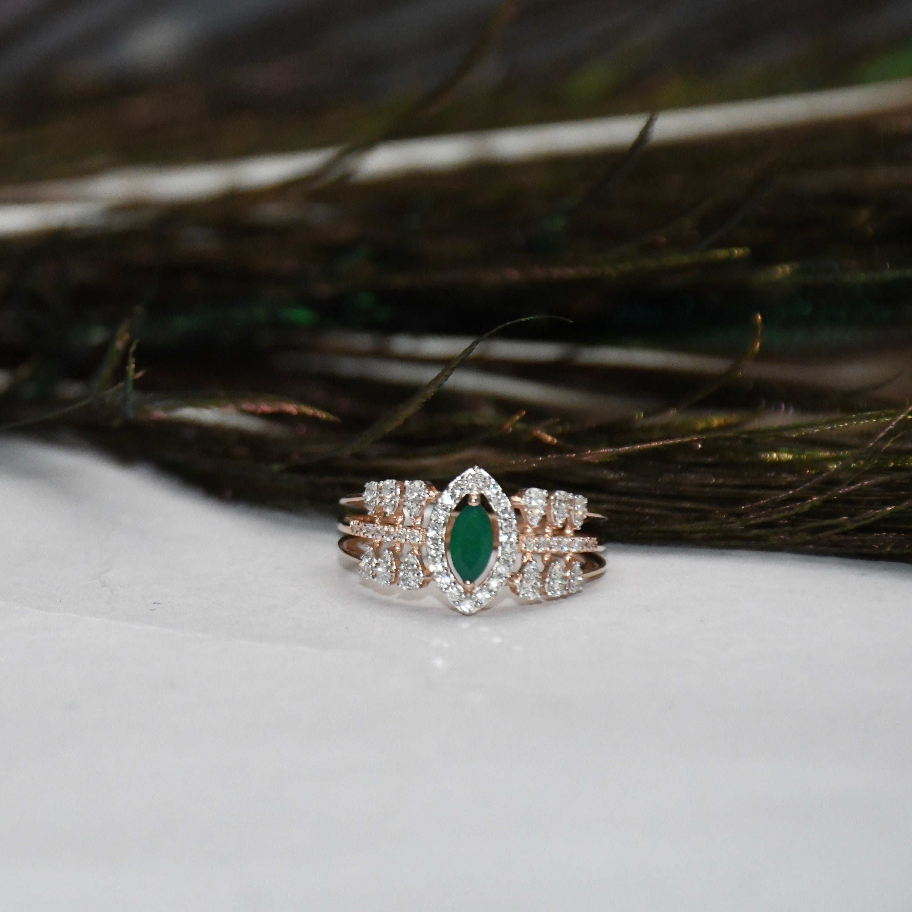 A rose gold ring with a marquise-cut green emerald center stone and surrounding pavé diamonds, displayed on a white surface with dark feathers in the background.