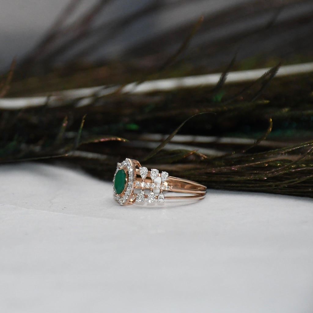 A rose gold ring featuring a green oval emerald center stone and diamond accents, resting on a white surface with dark decorative feathers in the background.