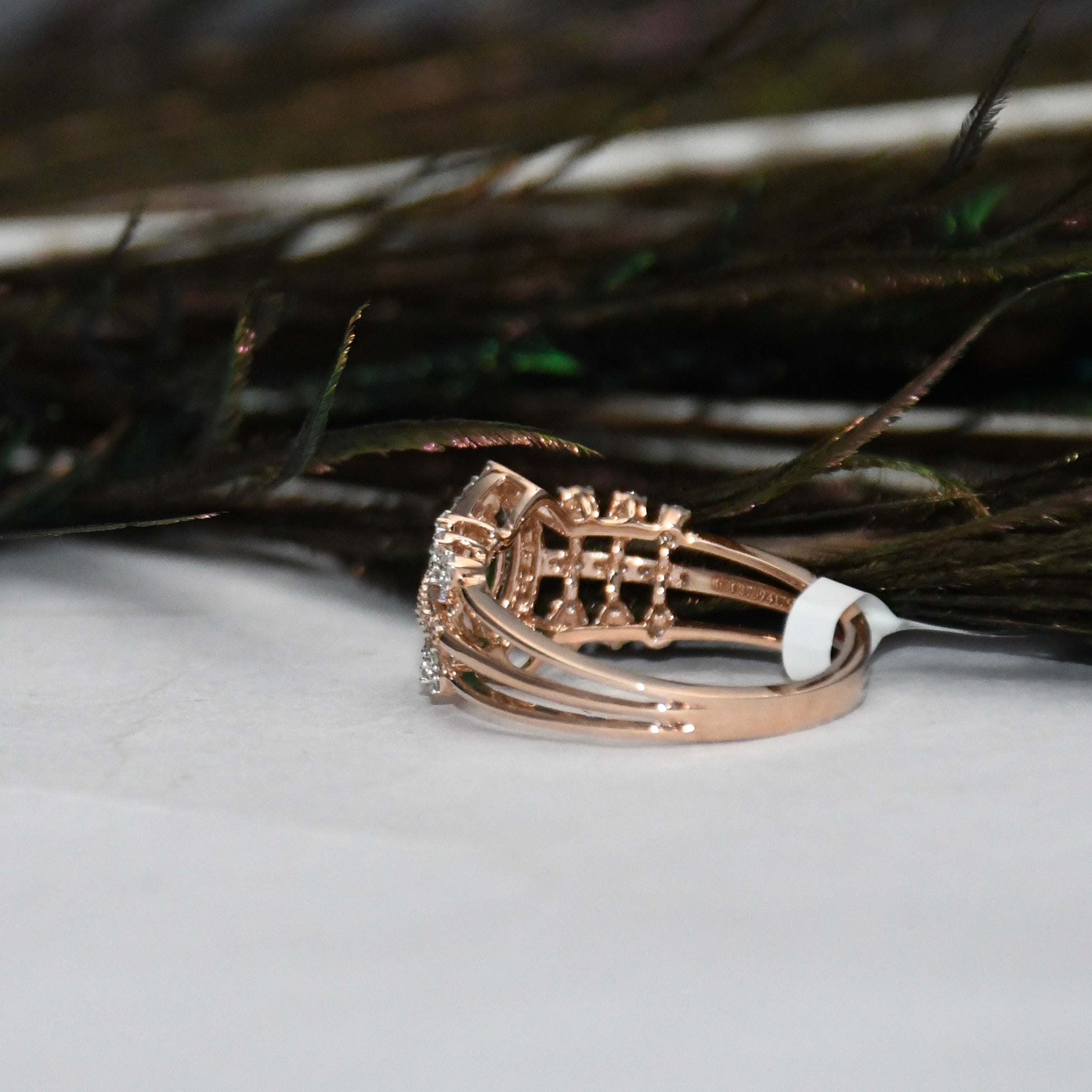 A rose gold ring with a basketweave diamond pattern, resting on a white surface with dark feathers in the background.