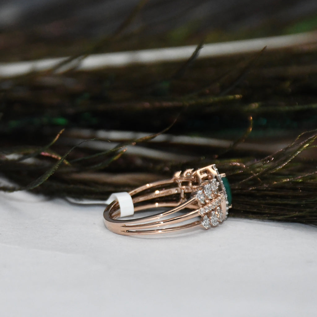 A rose gold ring with a cluster of clear stones and a green gemstone, resting on a white surface with dark feathers in the background.