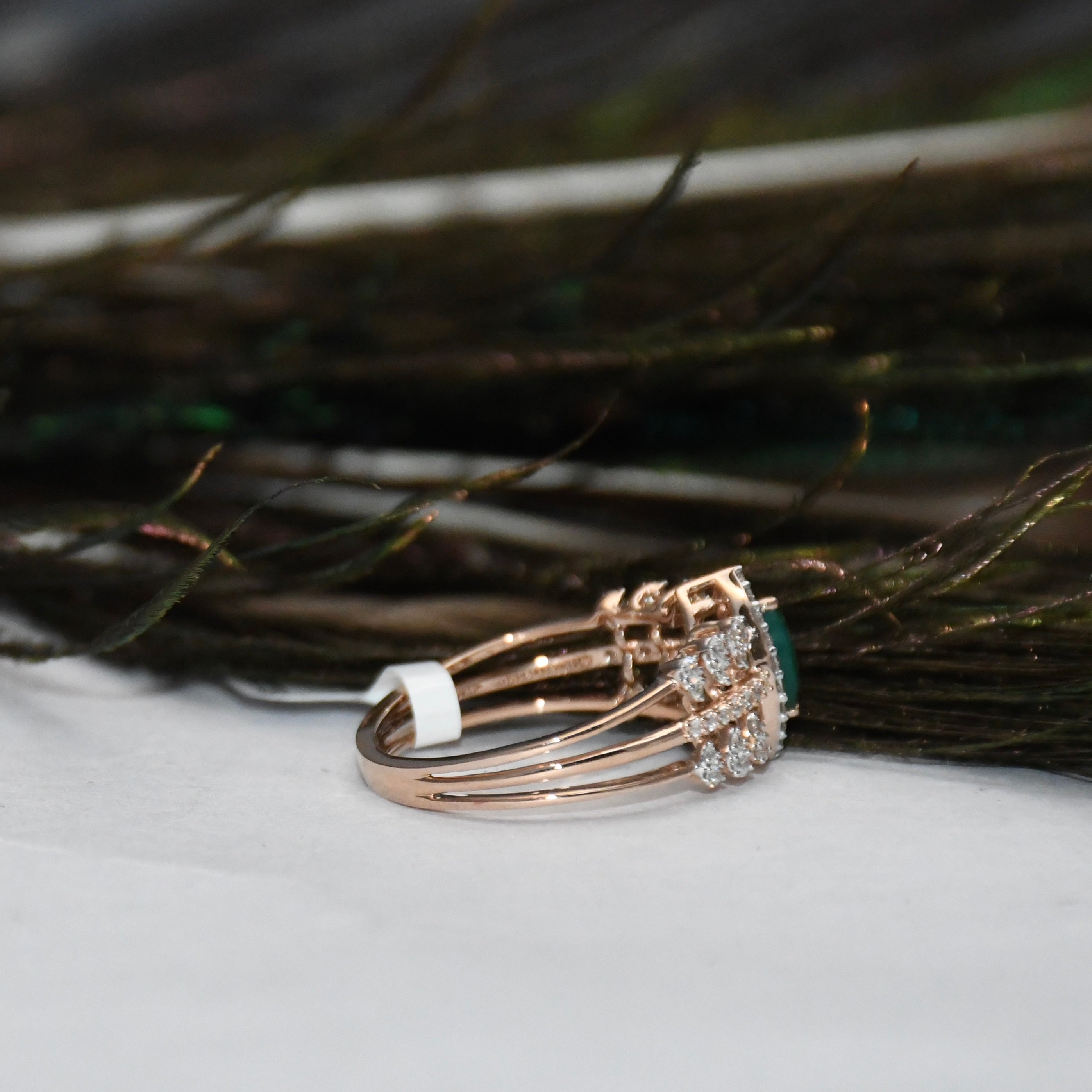 A rose gold ring with a cluster of clear stones and a green gemstone, resting on a white surface with dark feathers in the background.